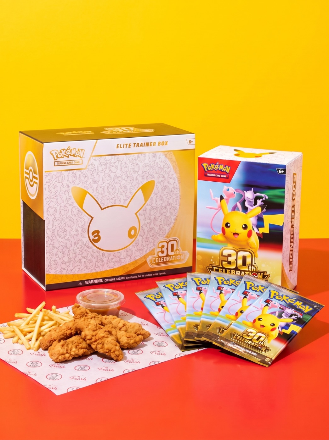 A Pokémon TCG 30th Celebration Elite Trainer Box and Booster Bundle with six booster packs beside a basket of Slim Chickens tenders